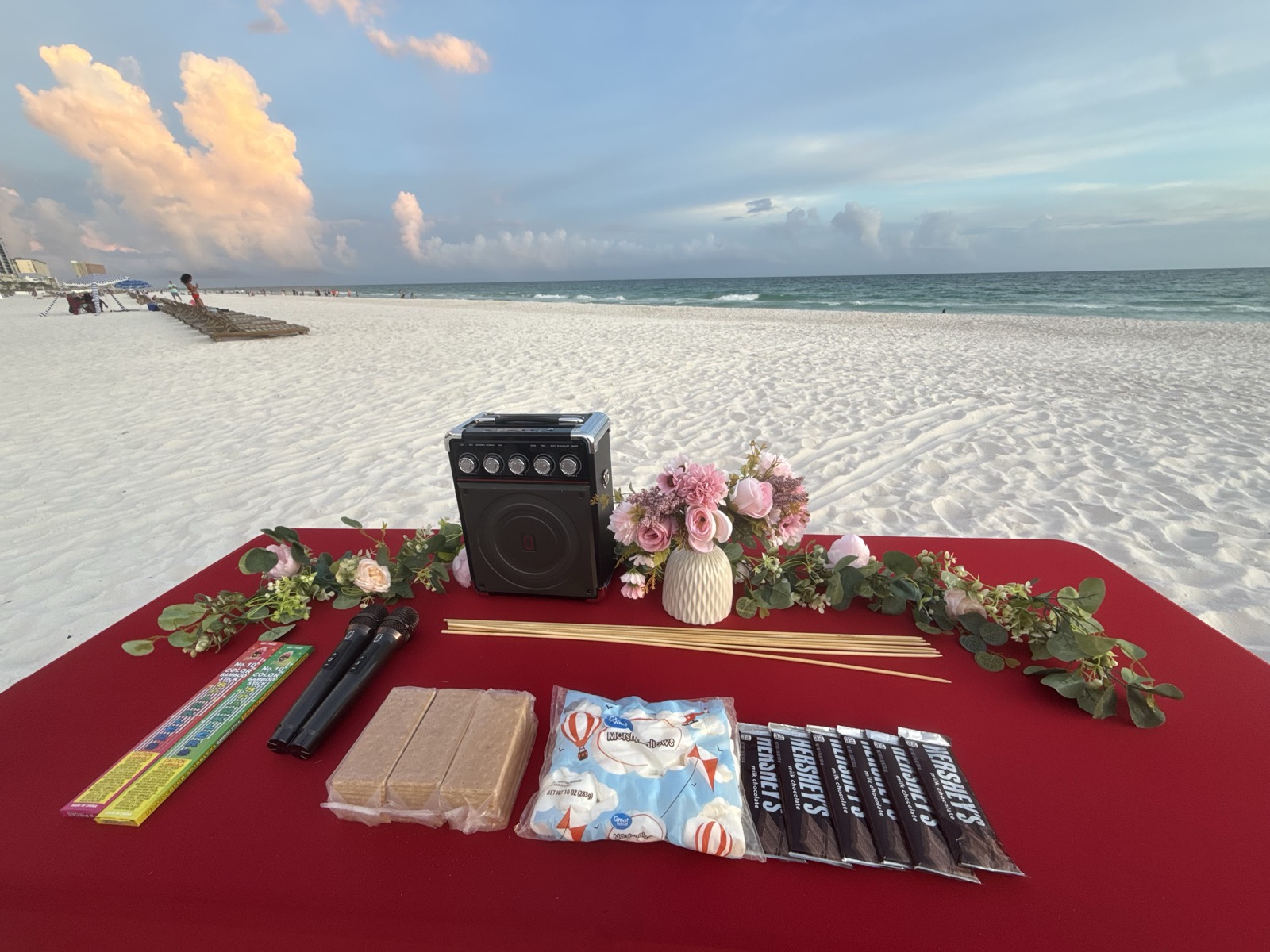 Glam Bonfire setup — red table with s'mores, sparklers, speaker, and florals on the beach